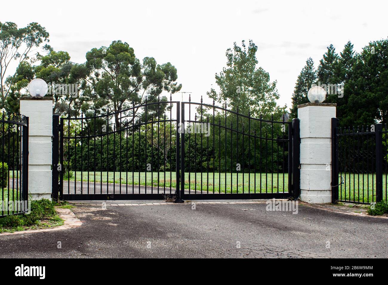 Black metal wrought iron driveway property entrance gates  set in brick fence, lights, green grass, garden trees Stock Photo