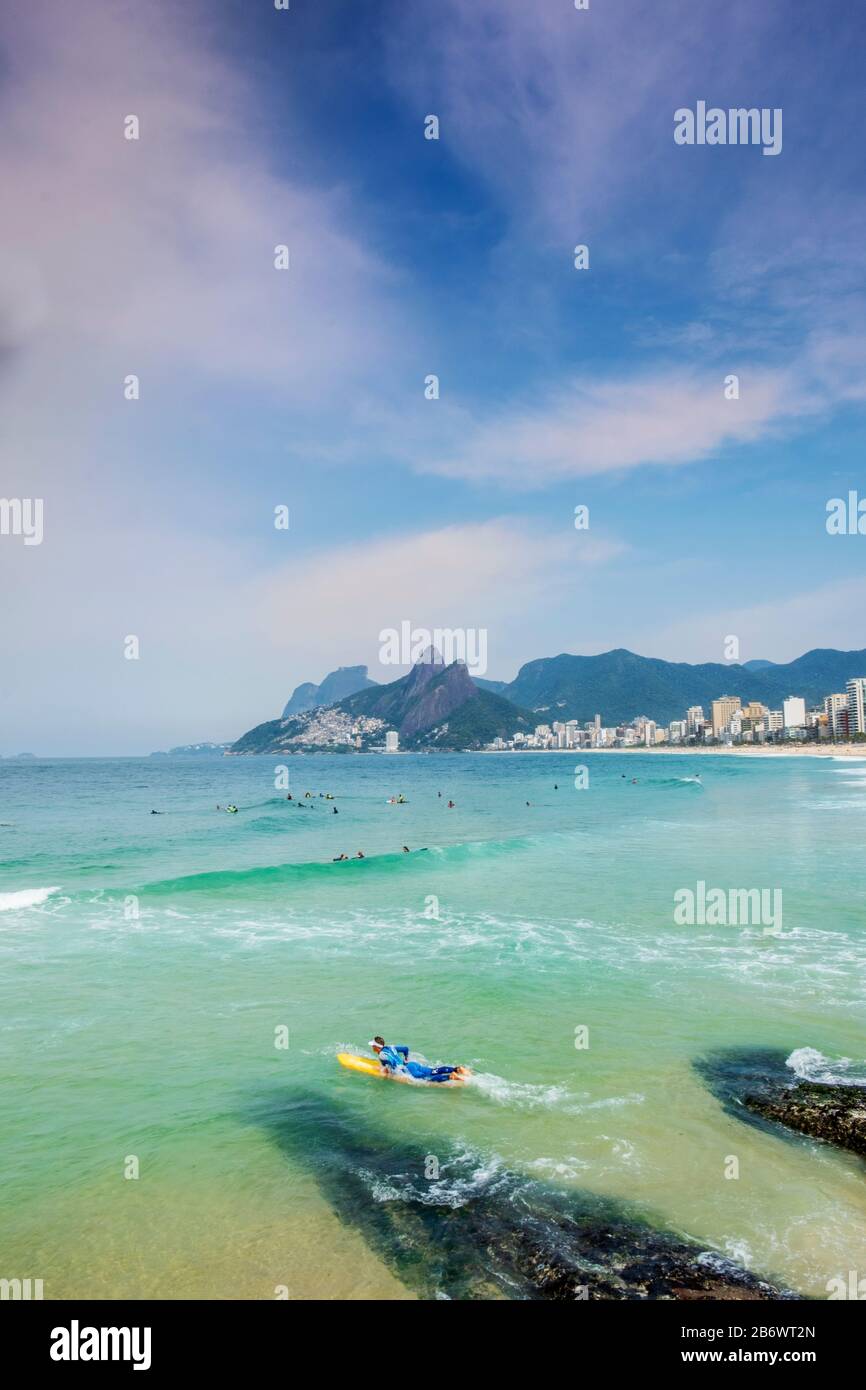 A male surfer entering the Atlantic Ocean at the Arpoador rocks with Ipanema beach, neighbourhood and the Dois Irmaos mountains in the background Stock Photo