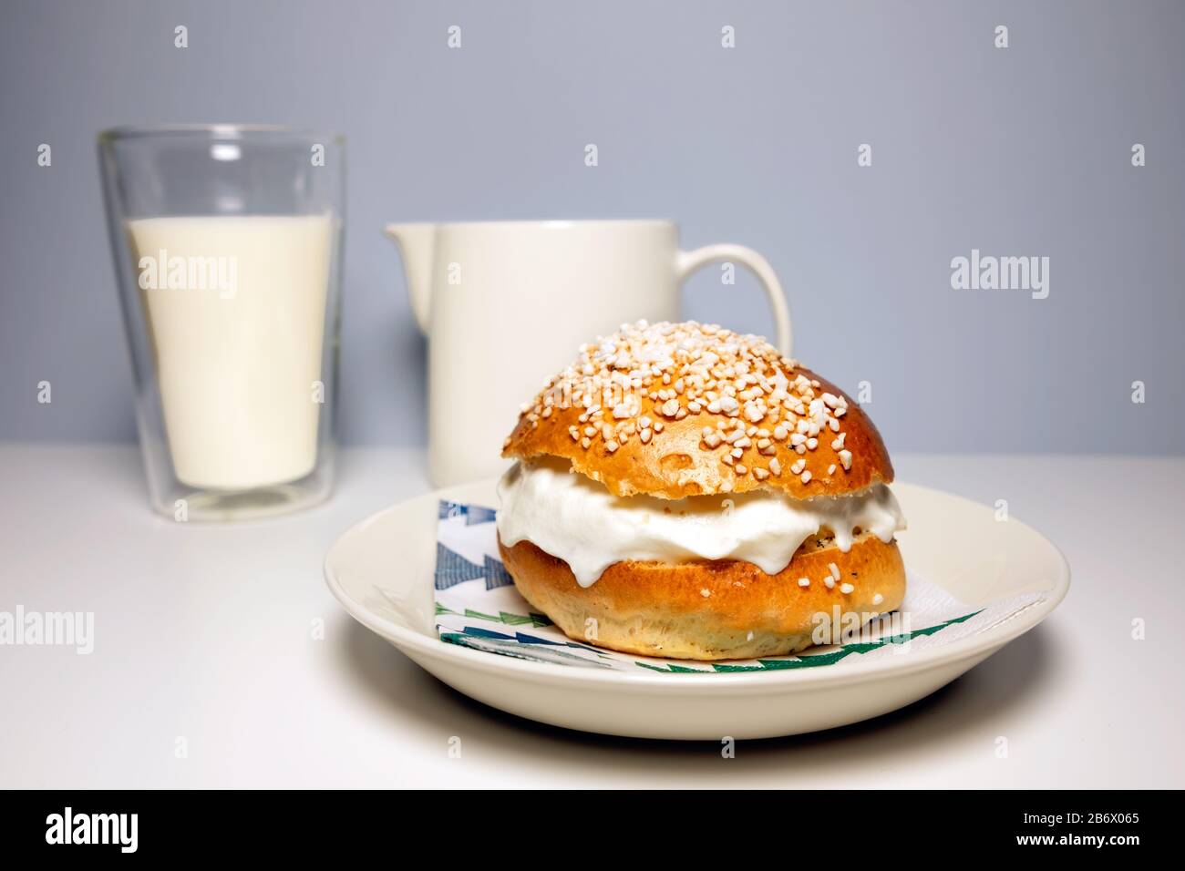 A traditional Scandinavian shrovetide bun with whipped cream, pearl sugar on top and a glass of milk on white table. Stock Photo