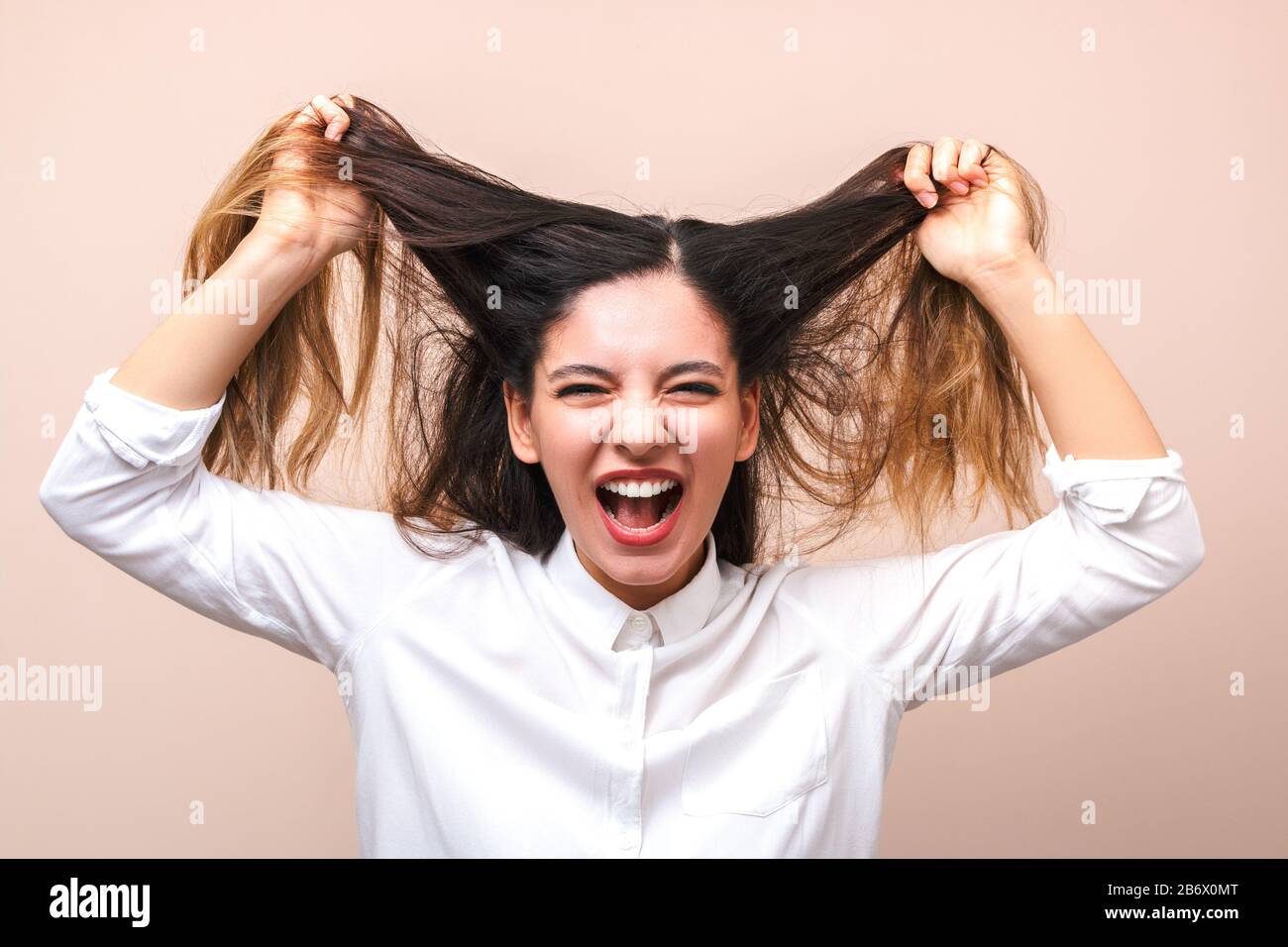 attractive brunette in white shirt pulls her hairs and screams against pink background. woman getting crazy Stock Photo