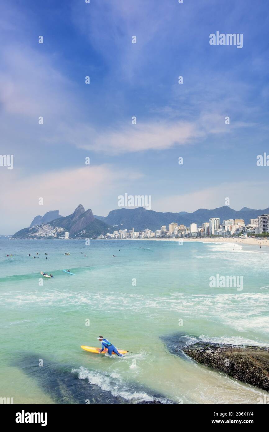 A male surfer entering the Atlantic Ocean at the Arpoador rocks with Ipanema beach, neighbourhood and the Dois Irmaos mountains in the background Stock Photo
