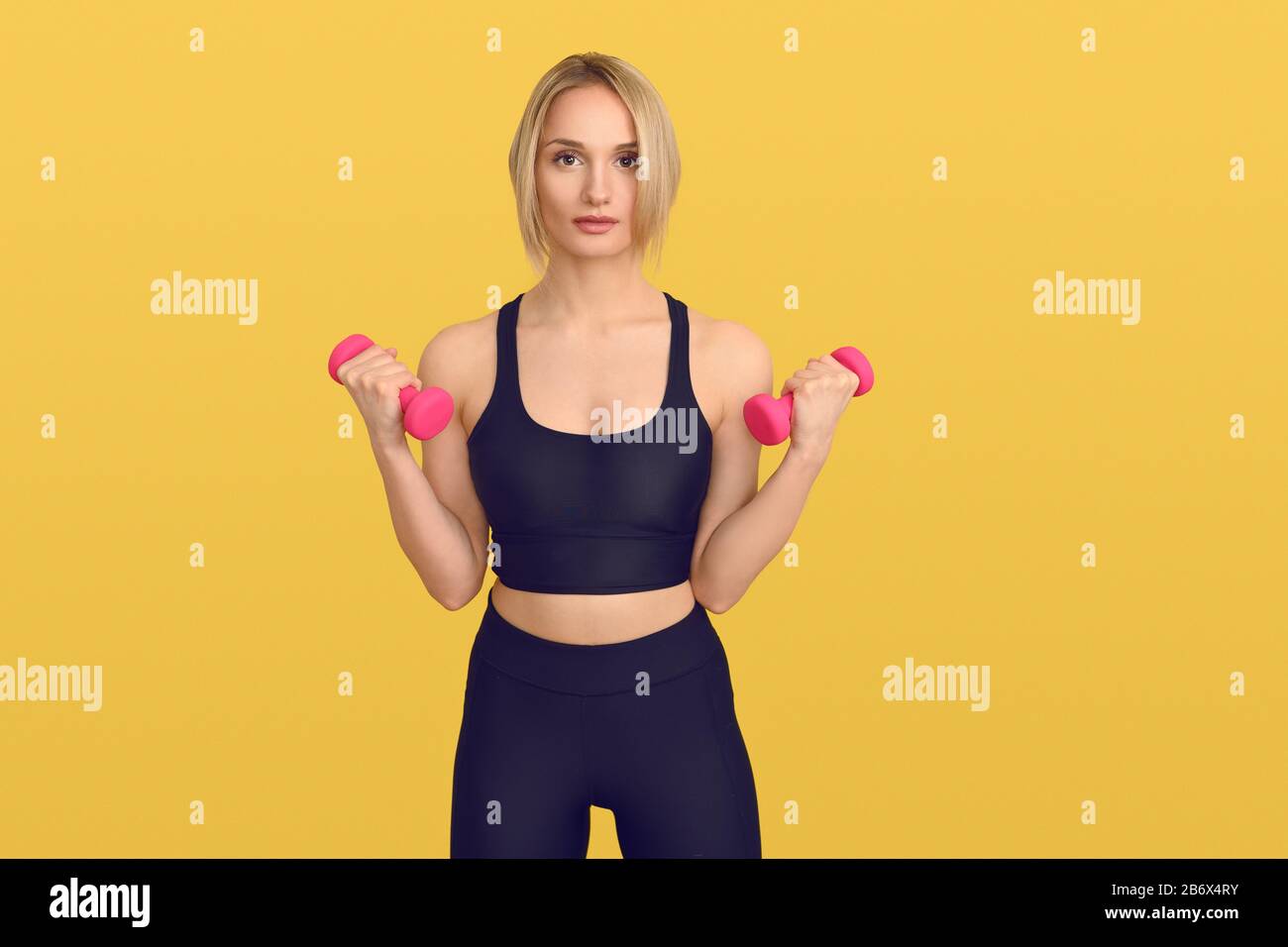 Pretty blond woman in black training top with small pink dumbbells in her hands, looking at camera. Close-up front portrait against plain yellow backg Stock Photo