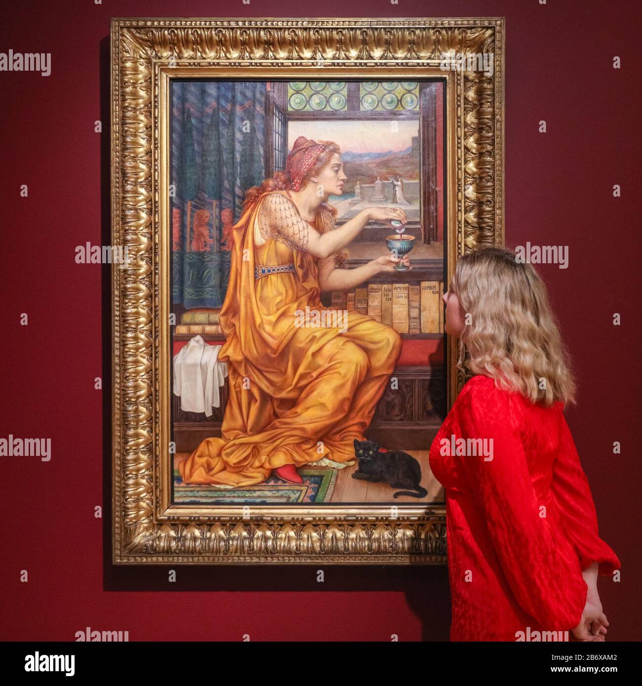 London, UK. 12th Mar, 2020. An assistant poses with Evelyn de Morgan's 'The Love Potion', depicting a woman in striking orange dress, 1903, oil on canvas. 'The Enchanted Interior' at Guildhall Art Gallery challenges the historic depiction of women in art with Pre-Raphaelite and contemporary female artists' works. The exhibition runs 13th March to 14th June, 2020. Credit: Imageplotter/Alamy Live News Stock Photo
