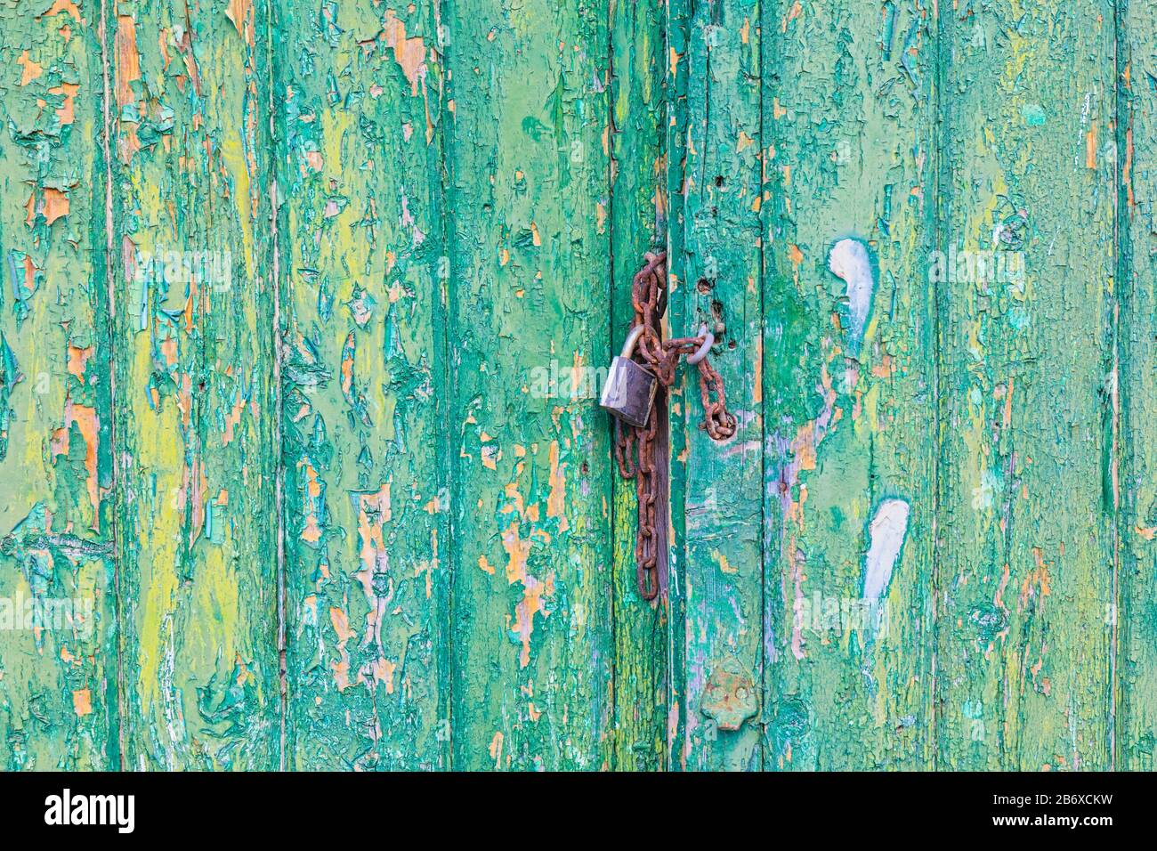 Rusty lock and chain holding weathered doors with flaking yellow and green paint closed. Stock Photo