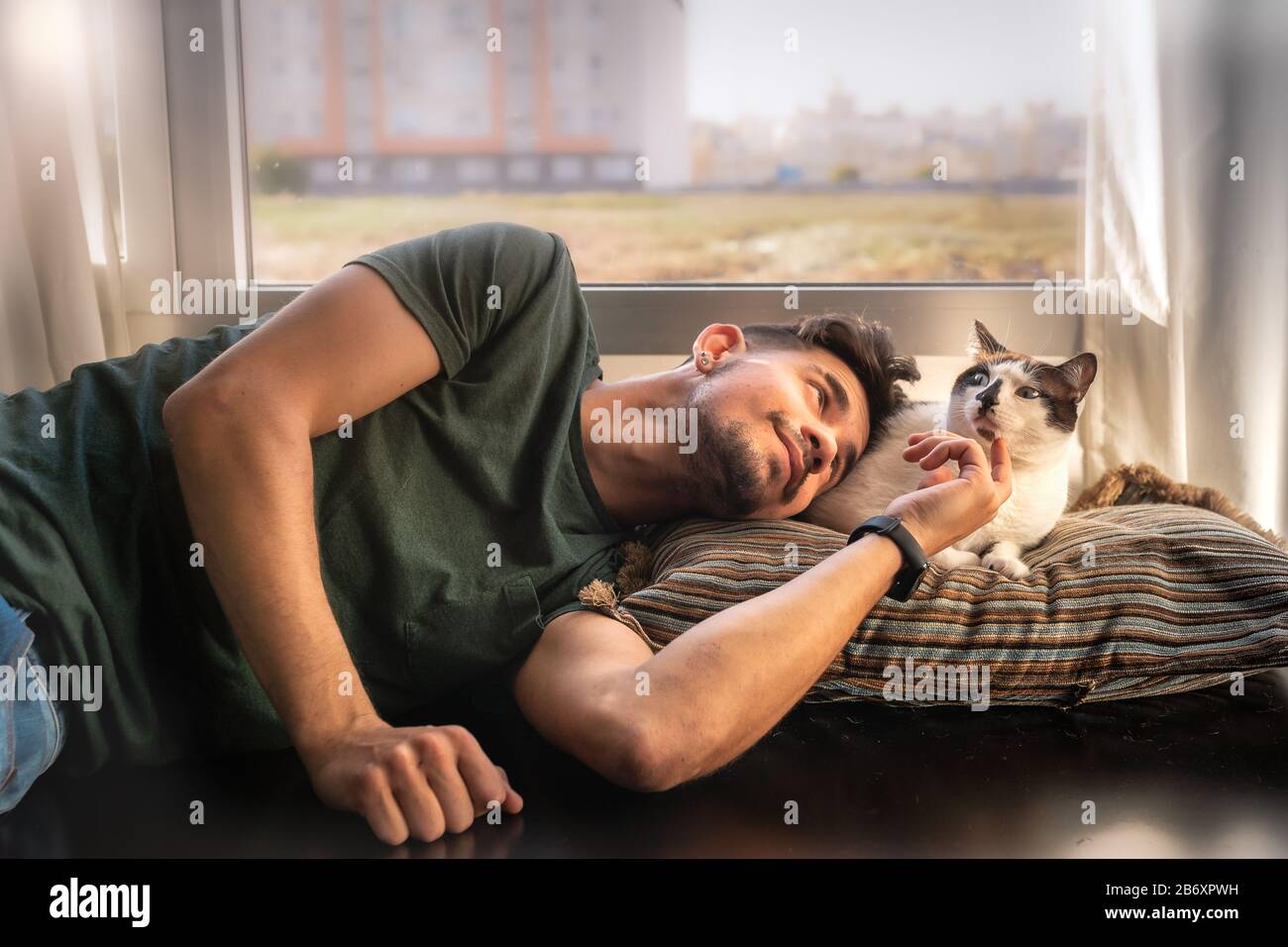 young man lying down lies next to a blue-eyed black and white cat and strokes his face Stock Photo