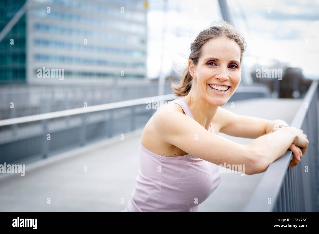 Happy young woman rests after jogging in the city and leans on a railing. Glückliche junge Frau ruht nach dem Joggen und stützt sich auf ein Geländer. Stock Photo