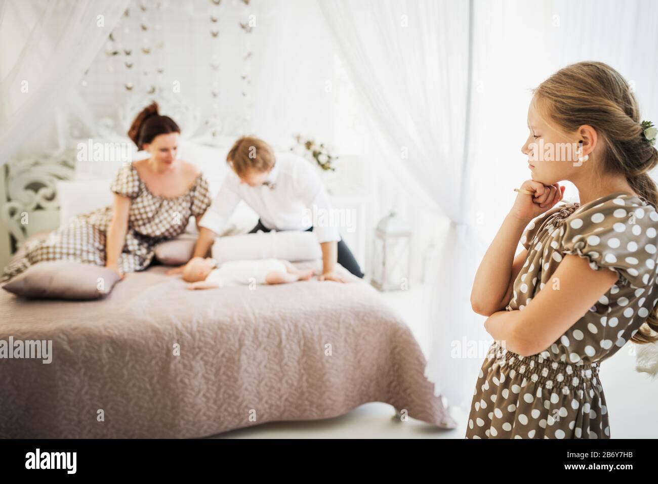 Little cute girl is jealous of her newborn brother lying on the bed next to the blurry happy parents. Concept of selfishness and the big difference in Stock Photo