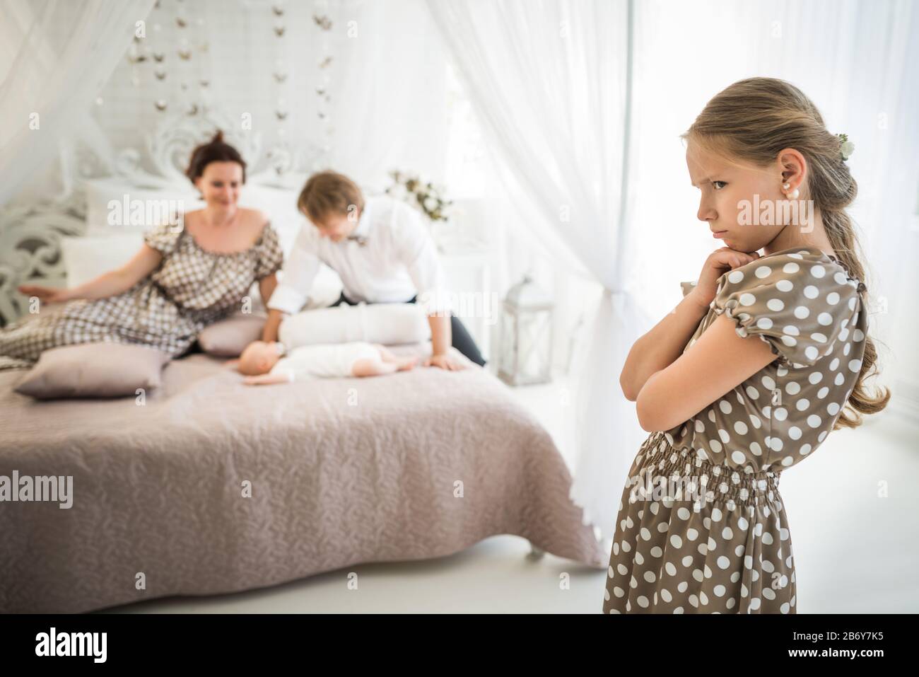 Little cute girl is jealous of her newborn brother lying on the bed next to the blurry happy parents. Concept of selfishness and the big difference in Stock Photo