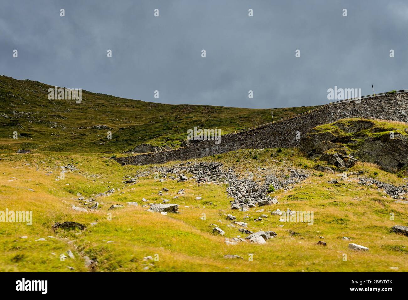 Upwards view of a mountain road railing with fallen rocks in the foreground and dark sky above Stock Photo