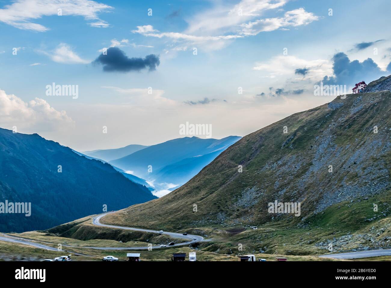 Mountain rescue house at the top of the mountain with cars passing on curvy road at the bottom and foggy forest covered mountains in the background Stock Photo