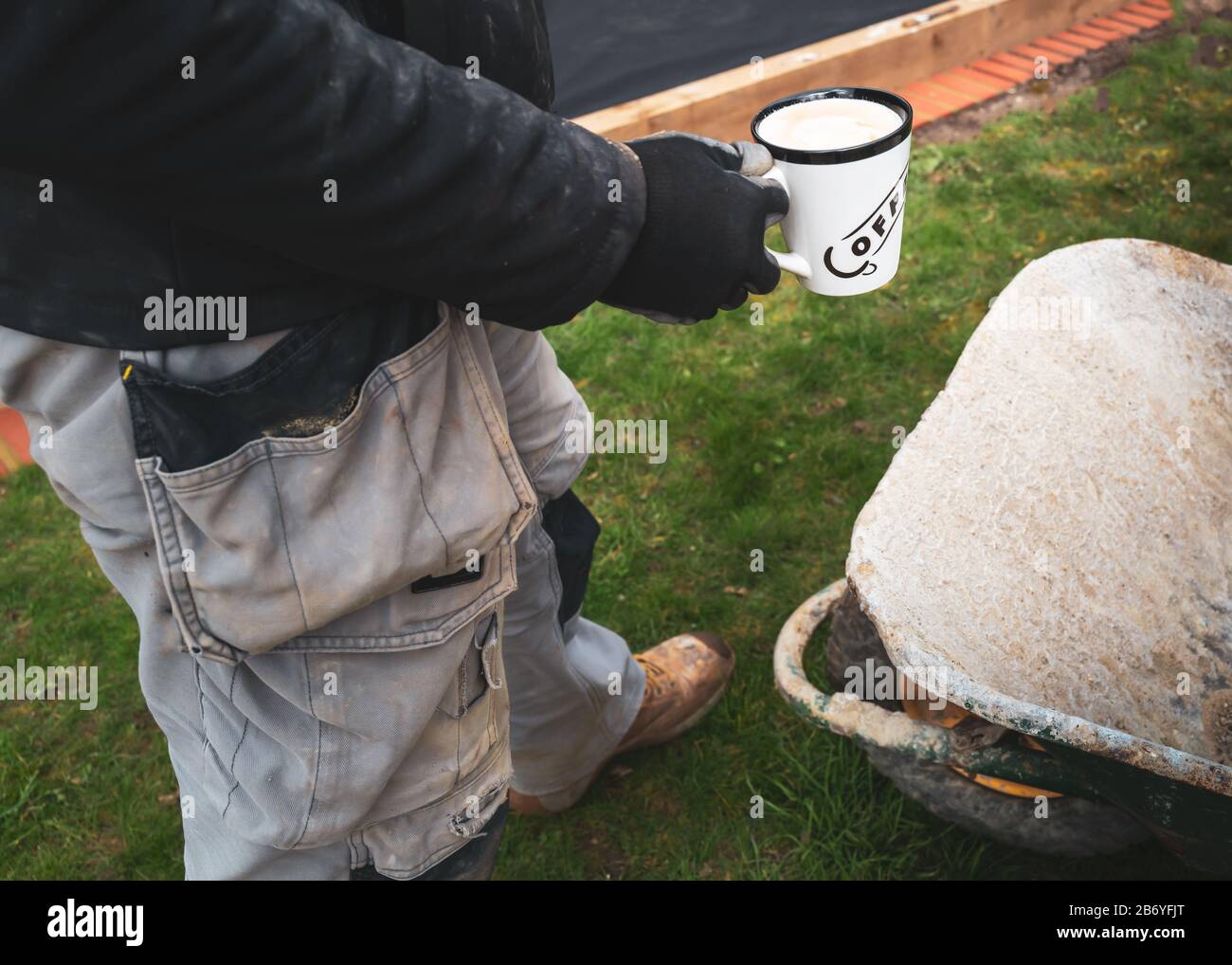 A cup of milky coffee being held by a workman in overalls by a wheelbarrow standing on grass in a garden or back yard. He is taking a break. Stock Photo
