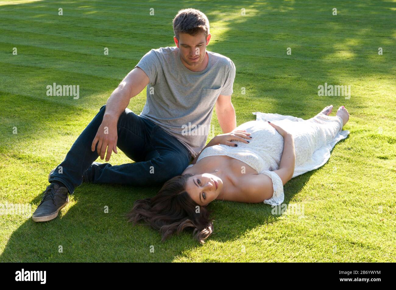 Beautiful pregnant young woman, wearing long white dress, lying down on the grass with her partner sitting next to her Stock Photo