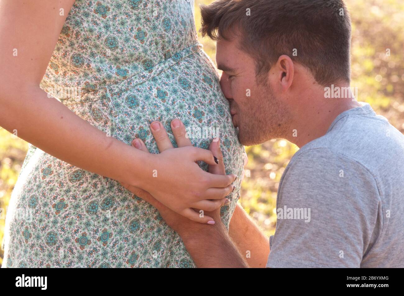 Pregnant young woman standing in a field, with her partner kneeling caressing her tummy, close-up Stock Photo