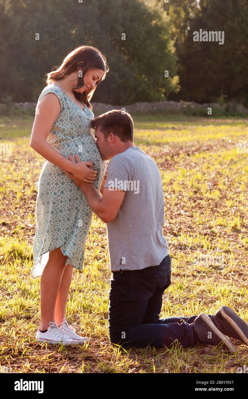 Pregnant young woman standing in a field, with her partner kneeling caressing her tummy Stock Photo