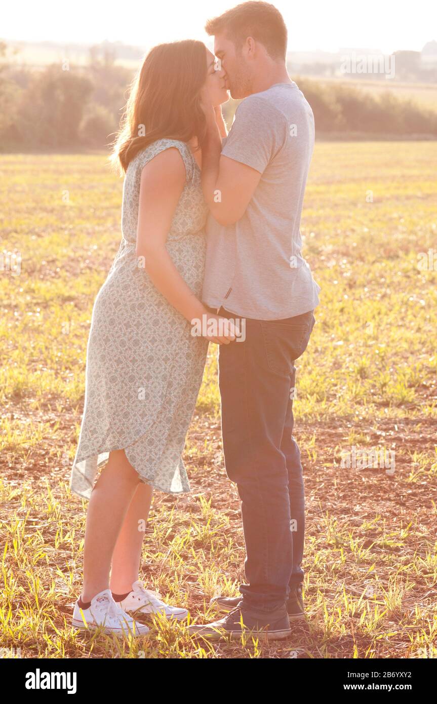 Pregnant young woman with her partner kissing and cuddling, standing in a field on a beautiful summers day Stock Photo