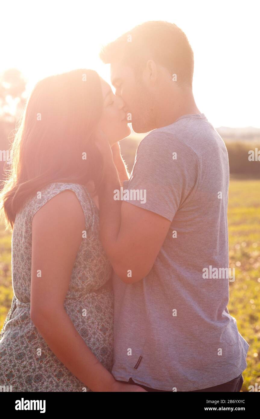 Pregnant young woman with her partner kissing and cuddling, standing in a field on a beautiful summers day Stock Photo