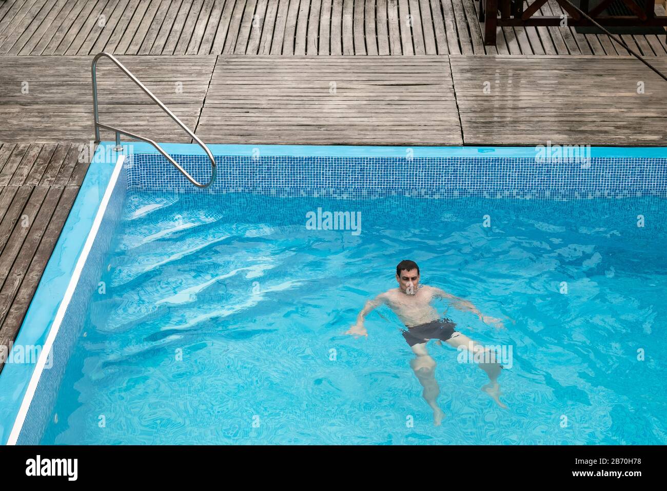 Young man swims in the blue pool outdoor at the territory of hotel. White guy of athletic build is in the water from afar. Tourism, vacation concept Stock Photo