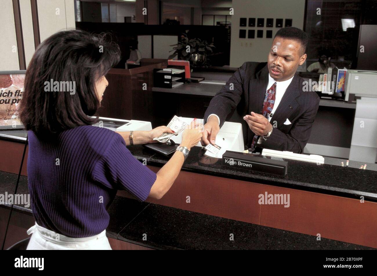  A bank teller wearing a suit and tie is talking to a customer while sitting at a desk in a bank.