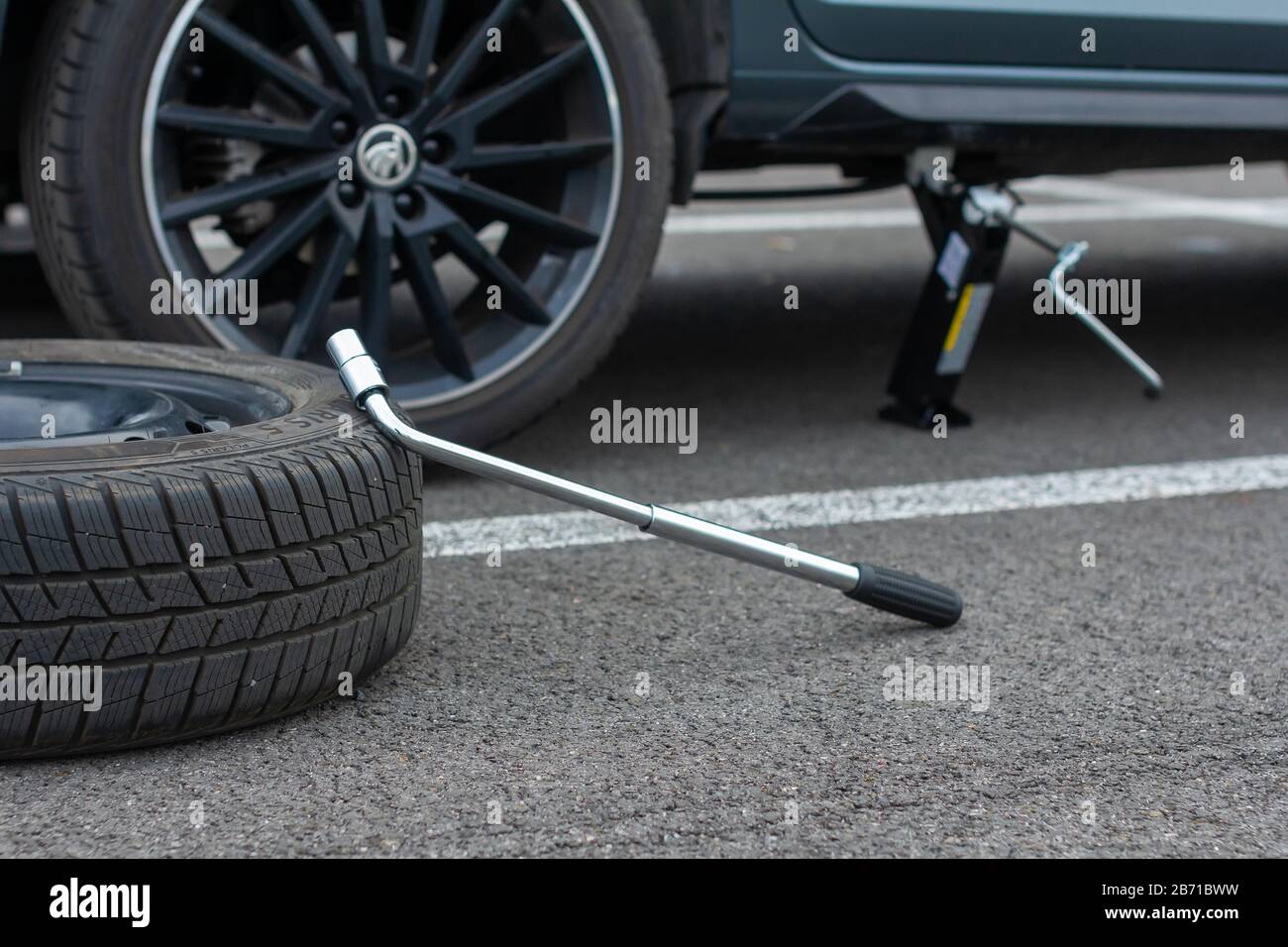 A flat tire car wheel and screwdriver are on a asphalt road on the broken Skoda Rapid background. Jack is lifting up a vehicle. Automobile service Stock Photo