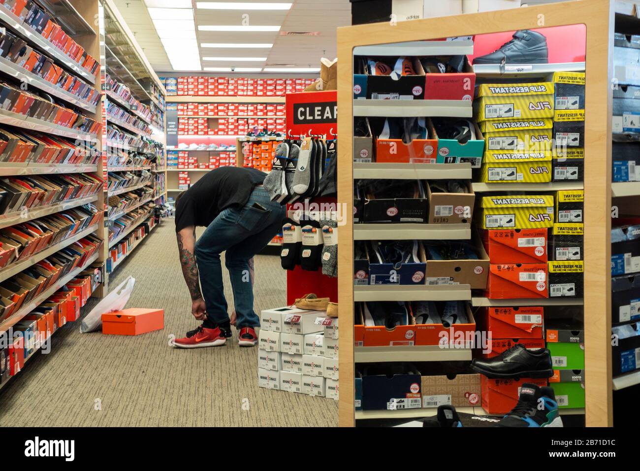 A 45-50 year old Caucasian tattooed man bends over trying on a pair of red athletic shoes inside a Famous Footwear store. USA. Stock Photo