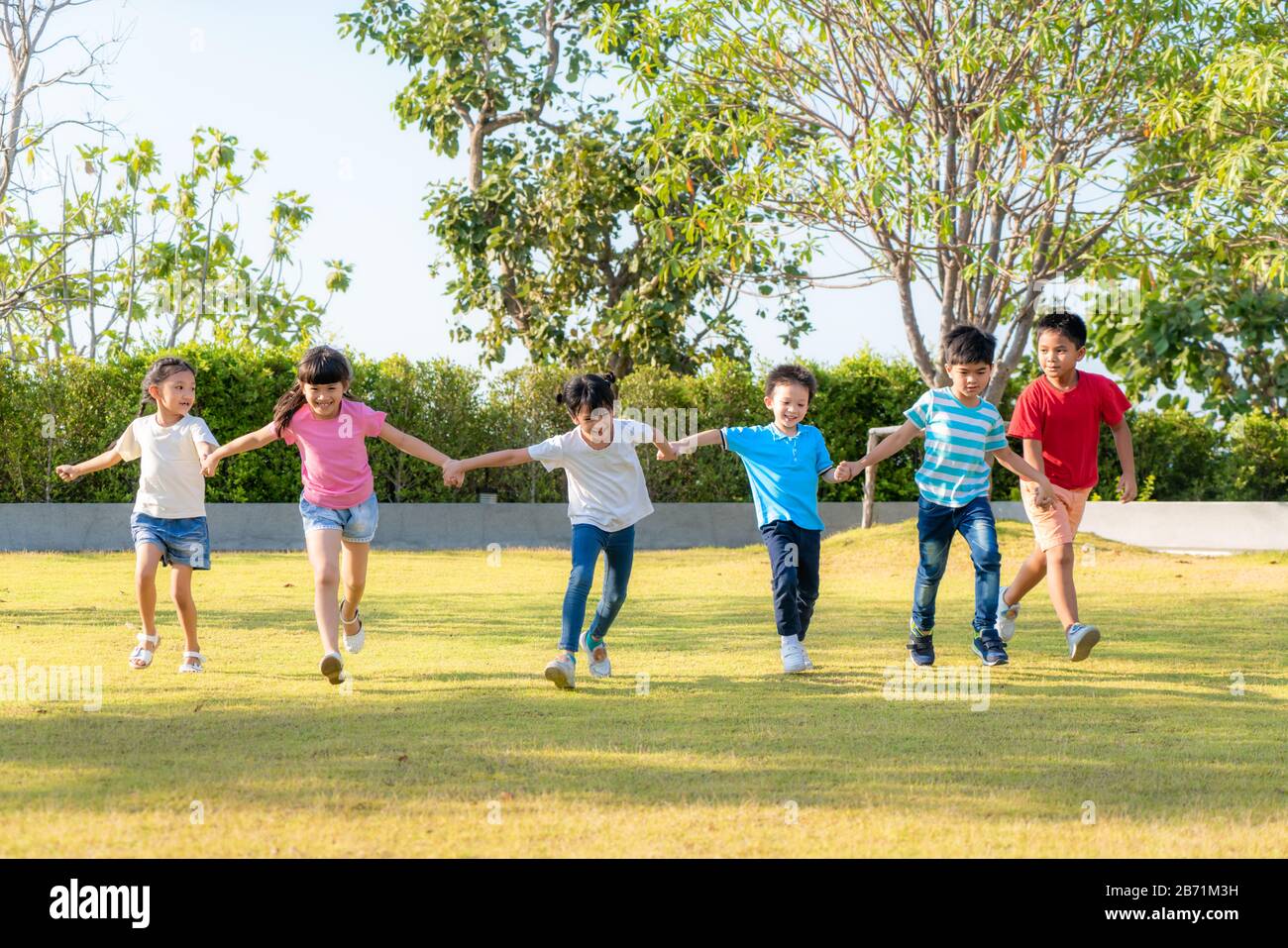 Large group of happy Asian smiling kindergarten kids friends holding hands playing and running in the park on sunny summer day in casual clothes. Stock Photo