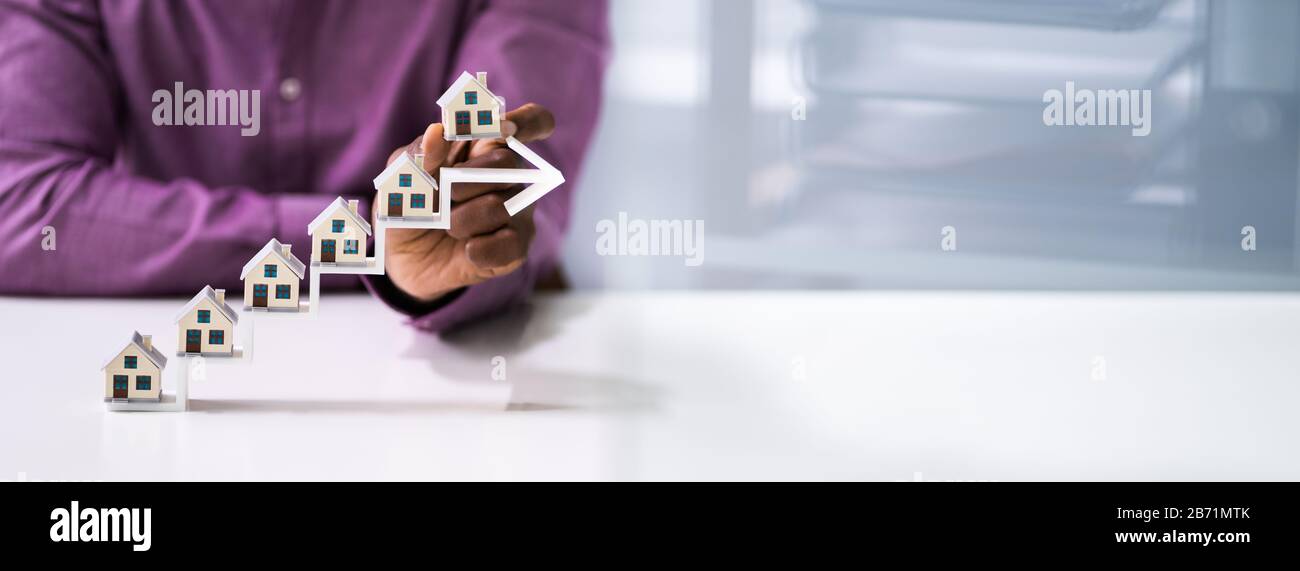 Close-up Of A Businessman's Hand Placing House Models On Increasing Blue Arrow Staircase Stock Photo
