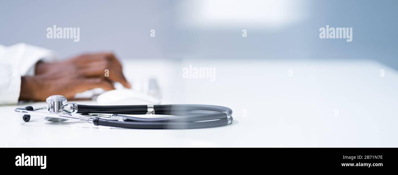 View Of Stethoscope With Doctor Using Computer Keyboard In Clinic Stock Photo