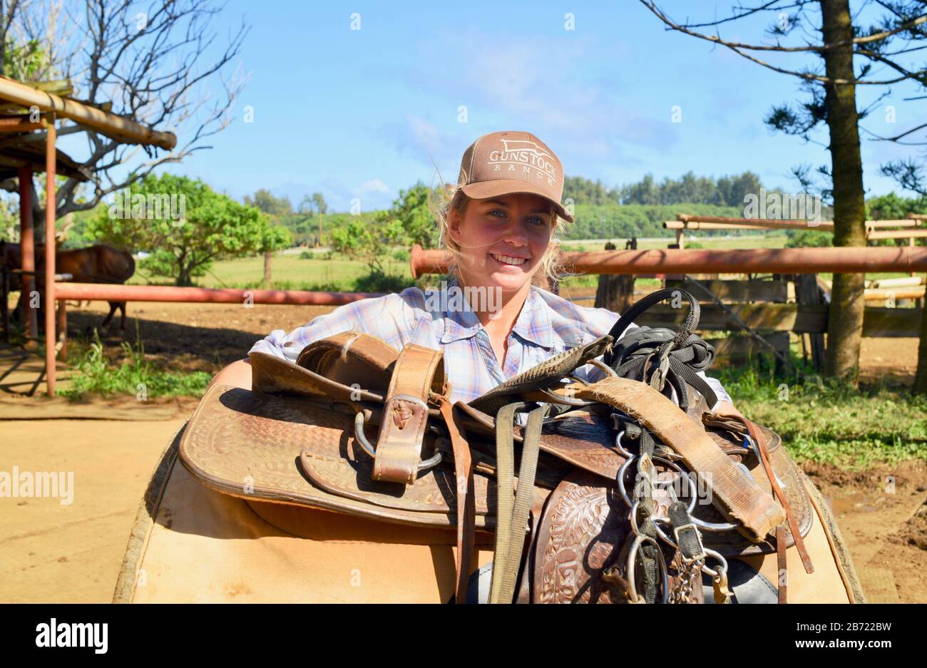 An attractive, young smiling cowgirl carrying a large, leather horse saddle at 900-acre Gunstock Ranch, Oahu Island, Laie, Hawaii, USA Stock Photo