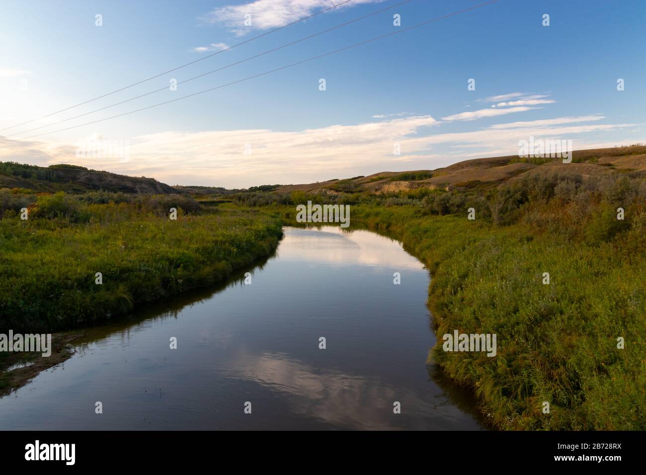 A calm and peaceful stream flowing through a valley in rural country side of the Saskatchewan Prairies in Canada with the sky reflecting in the water Stock Photo