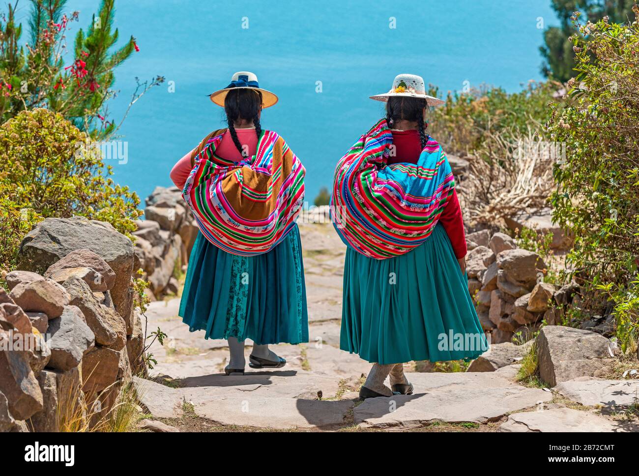 Two indigenous Quechua women in traditional clothes walking down the path to the harbor of Isla Taquile and the Titicaca Lake in the background, Peru. Stock Photo