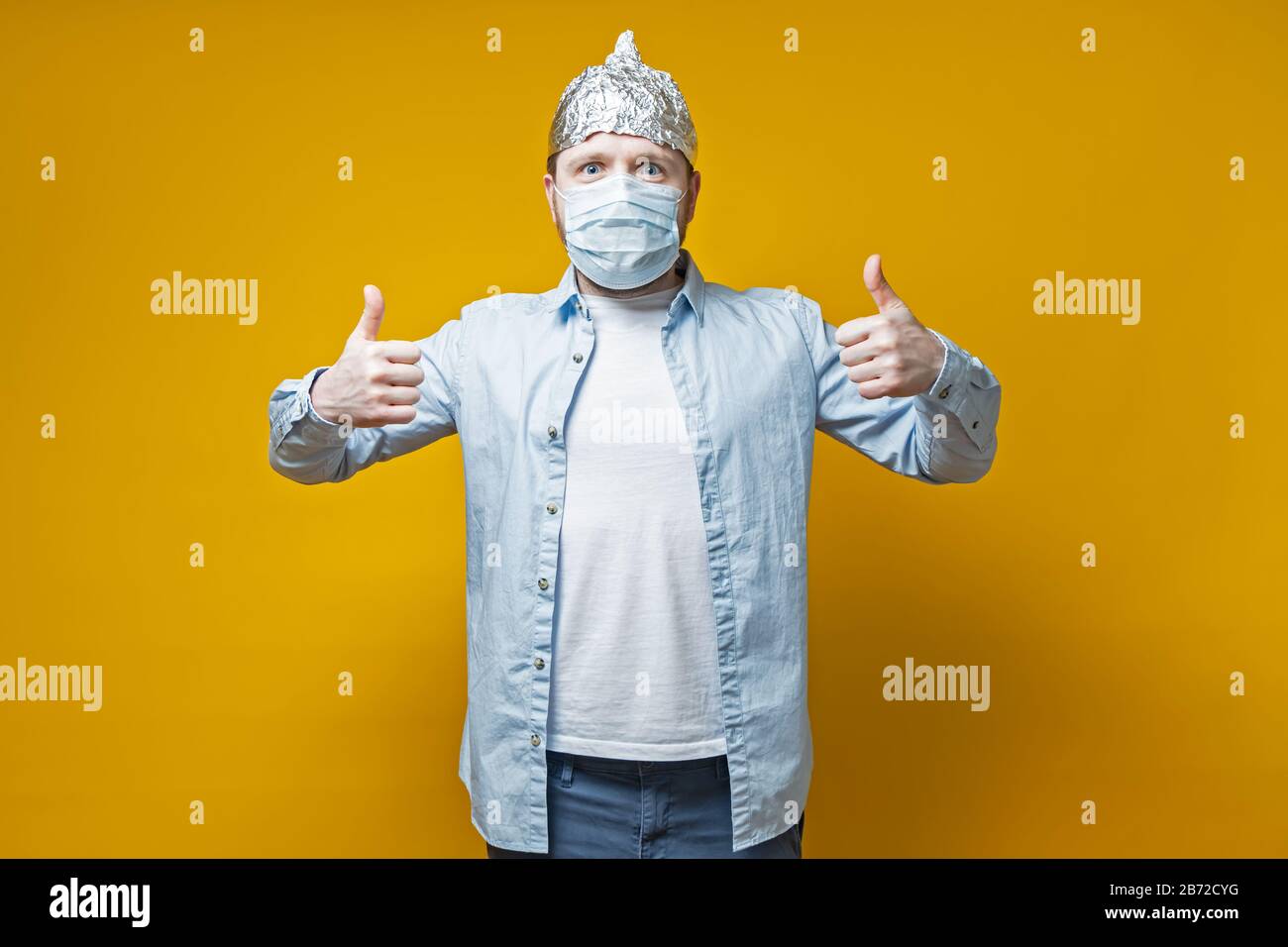 Man protected himself from the virus, put on a medical mask and a tin foil hat, he makes a thumbs up gesture and looks joyfully. Stock Photo