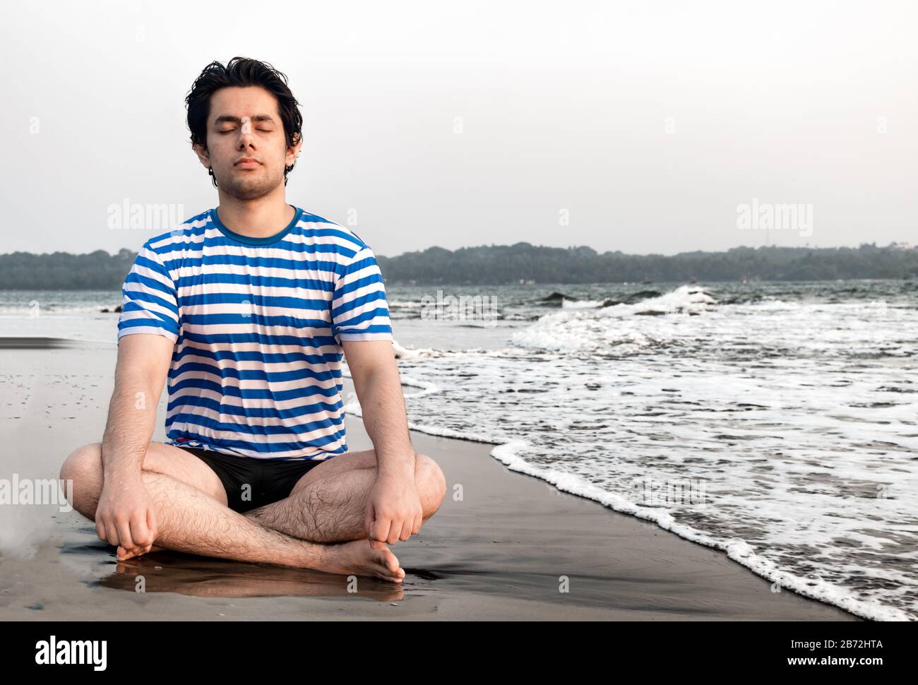 Handsome good looking boy in his early 20s, is meditating for physical and mental well being, while sitting in lotus position on beach. Yoga, Heath Stock Photo