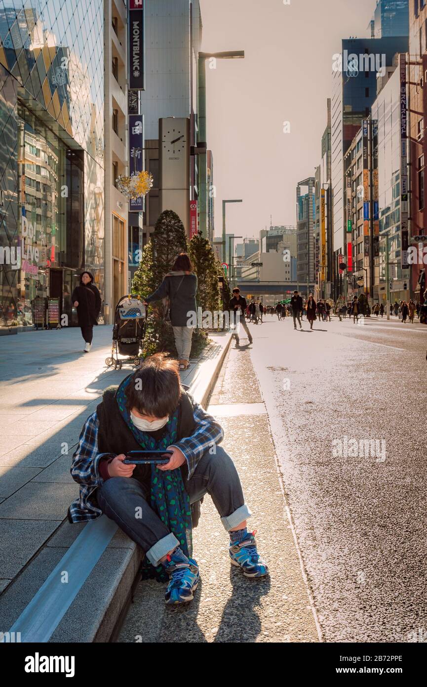 View down Shin juku in Tokyo, skyscrapers and shoppers in the background, as young boy, wearing a mask, sits on gutter playing his electronic device. Stock Photo