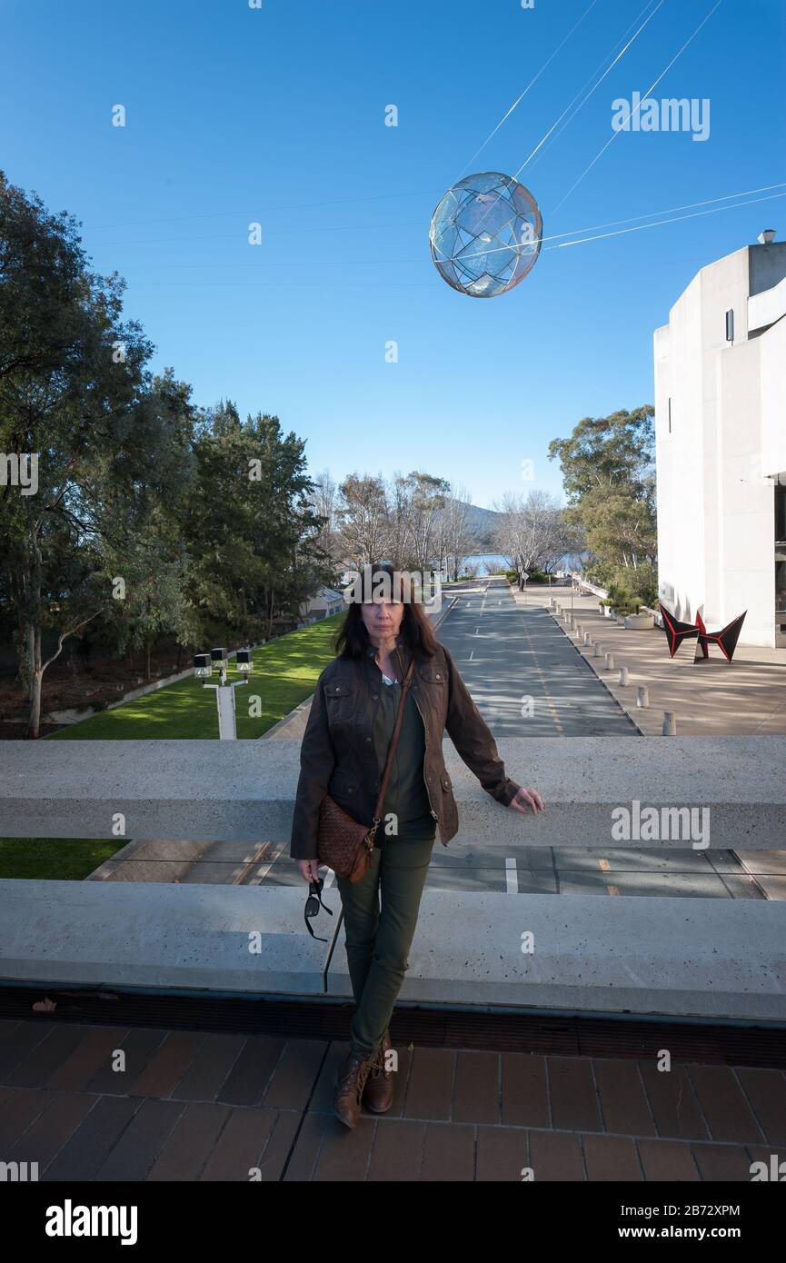 Female tourist dressed for winter standing on an elevated walkway across a Canberra road with a suspended spherical art installation overhead. Stock Photo