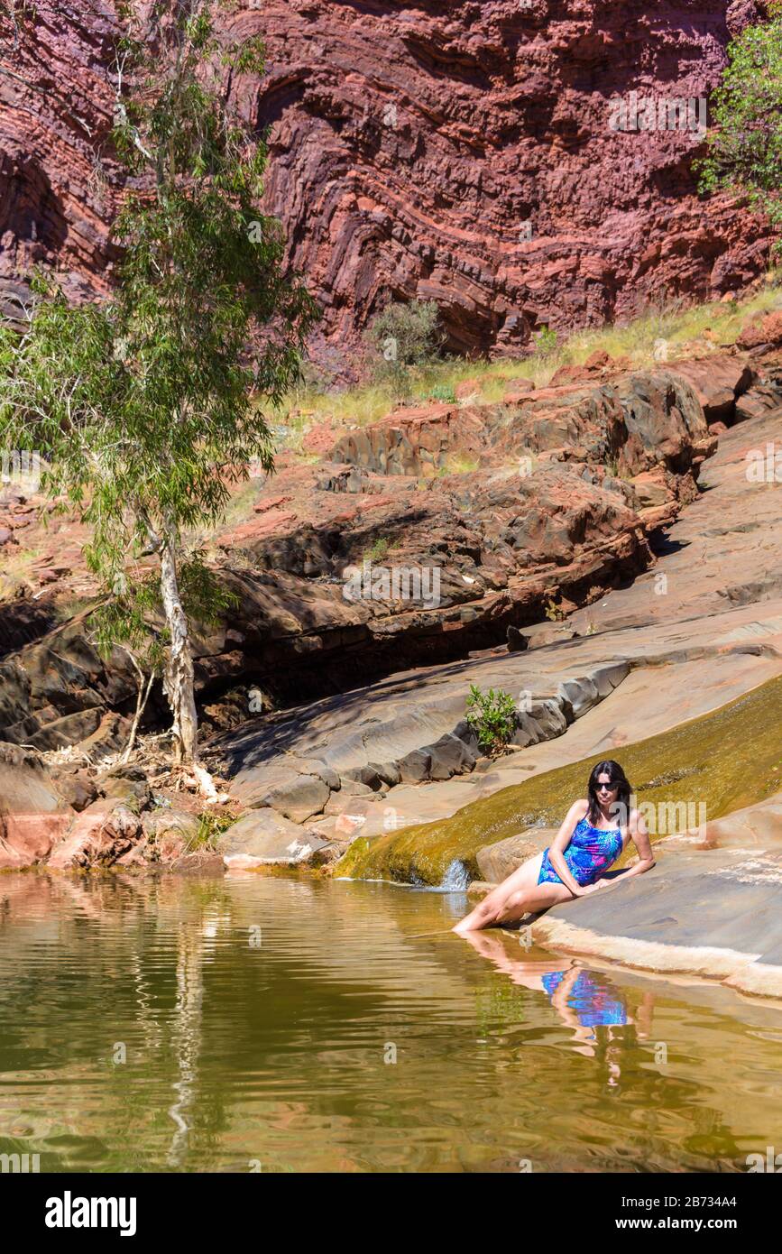 View of female tourist in bright blue swimsuit laying beside a crystal clear freshwater of the rugged Hamersley Gorge in Western Australia. Stock Photo