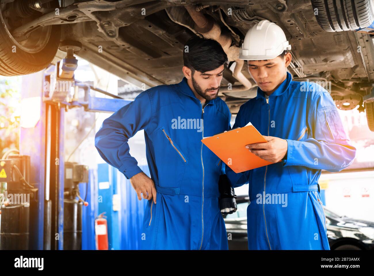 Mechanic in blue work wear uniform inspects the car bottom with his assistant. Automobile repairing service, Professional occupation teamwork. Stock Photo