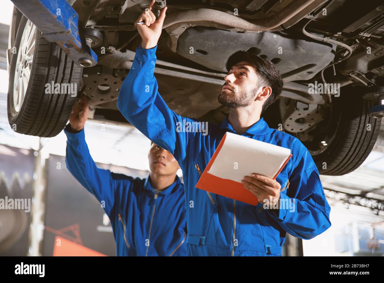 Mechanic in blue work wear uniform inspects the car bottom with his assistant. Automobile repairing service, Professional occupation teamwork. Stock Photo