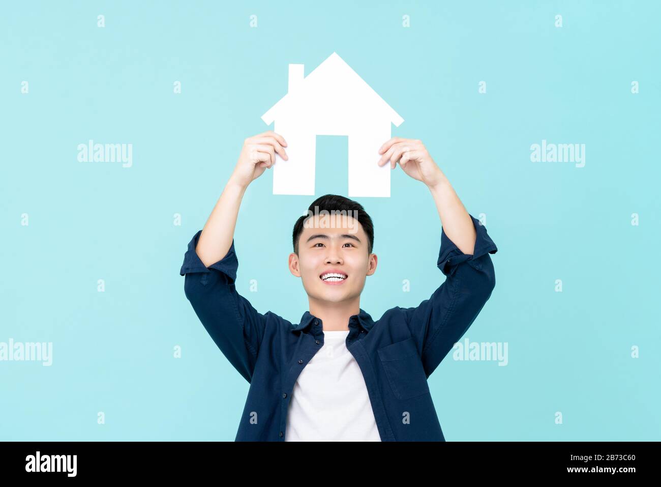 Happy young Asian man holding house sign overhead isolated on light blue background for real estate concepts Stock Photo