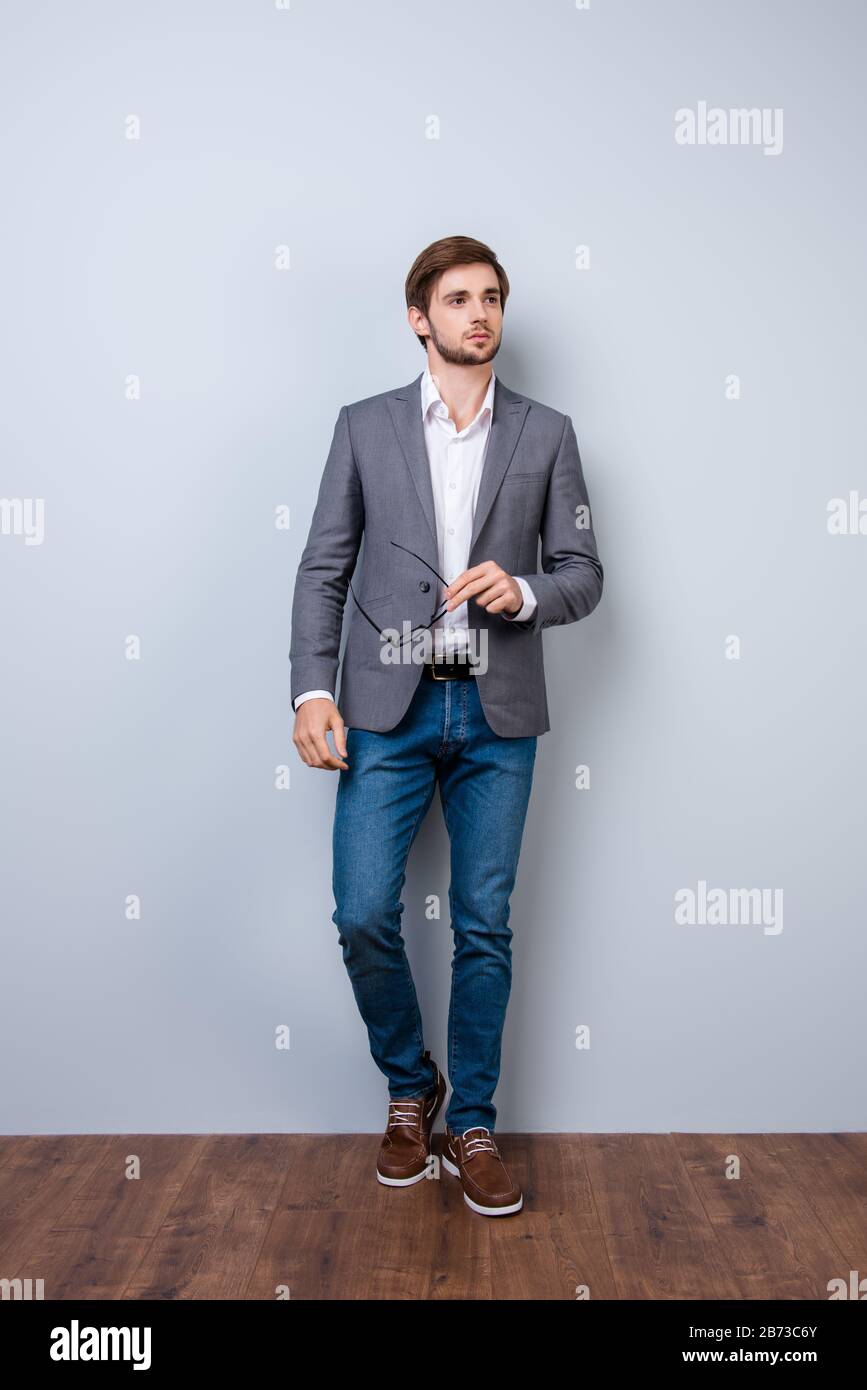 Full-length portrait of young handsome confident man in jacket holding glasses in his hand against gray background. Stock Photo