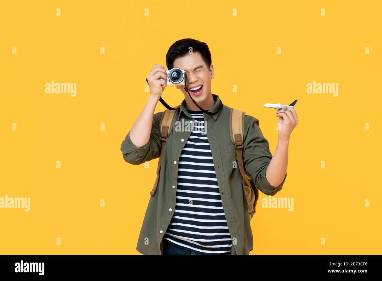 Young handsome smiling Asian tourist man holding plane model and camera isolated on yellow background Stock Photo