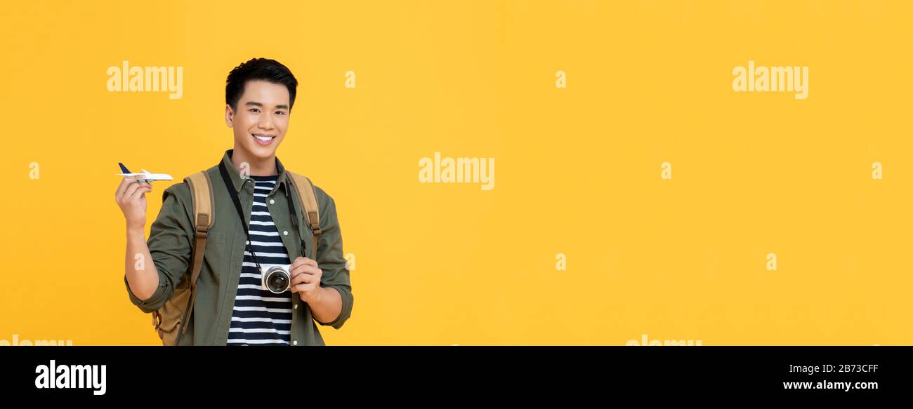Young handsome smiling Asian tourist man holding plane model and camera isolated on yellow banner background with copy space Stock Photo