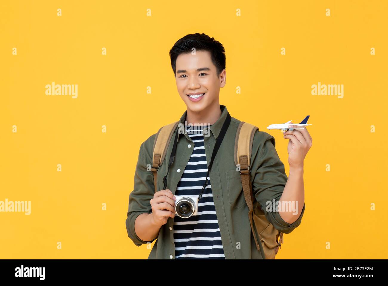 Young handsome smiling Asian tourist man holding plane model and camera isolated on yellow background Stock Photo