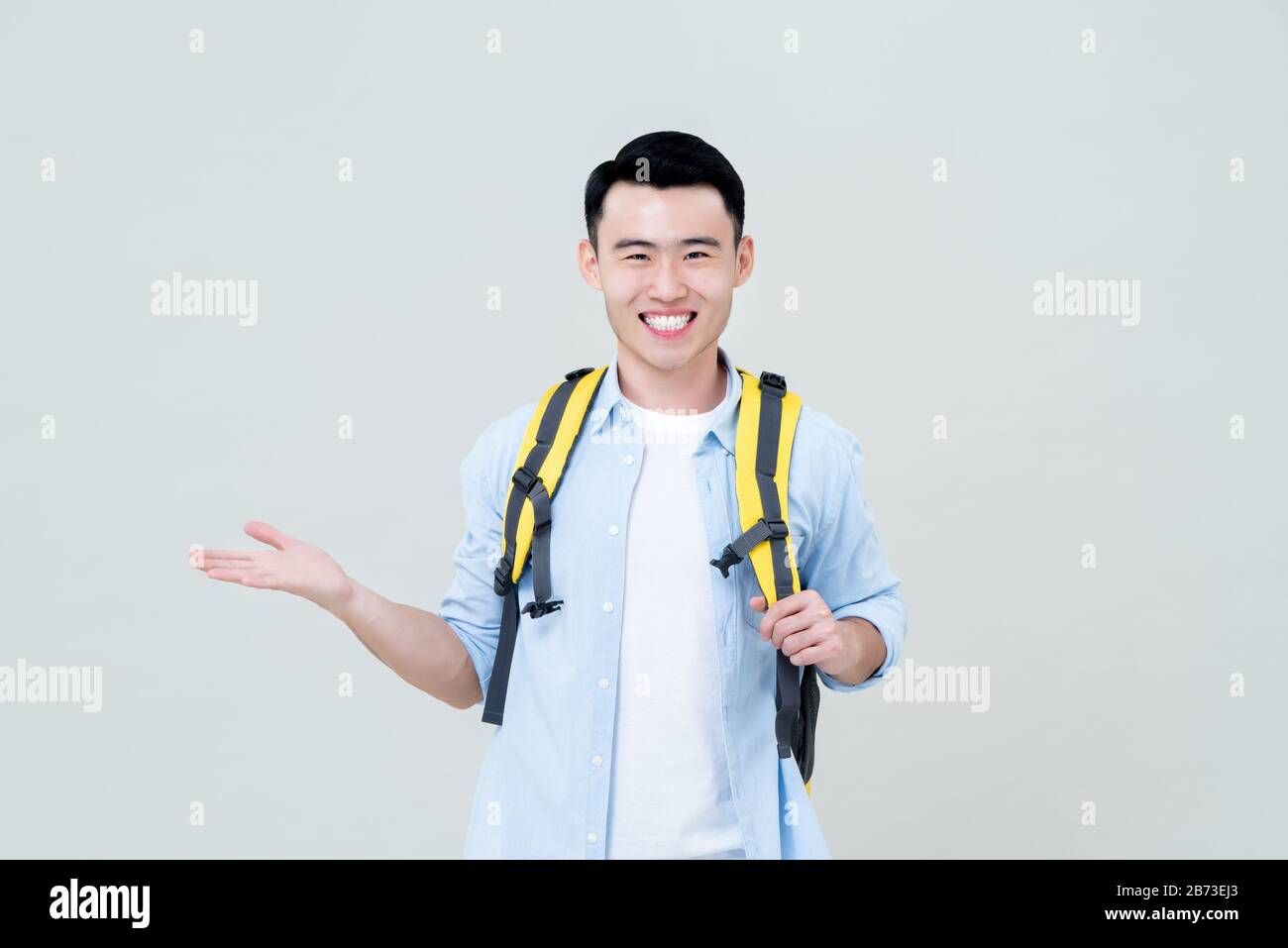 Waist up portrait of young confident smiling Asian tourist man backpacker doing  open palm gesture in gray studio background Stock Photo