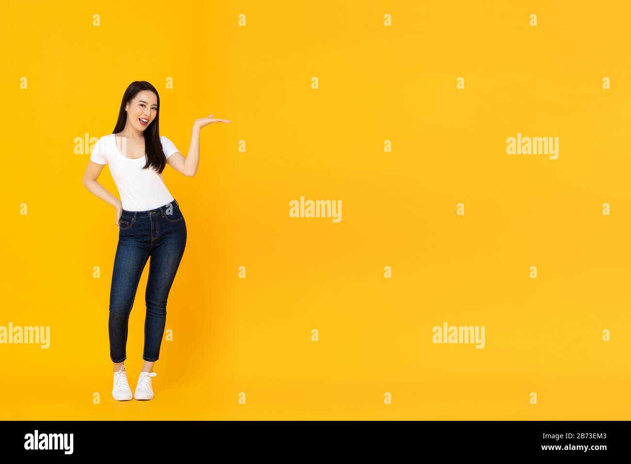 Full lenght portrait of smiling confident young beautiful Asian female model doing open palm gesture to empty space beside in yellow isolated studio b Stock Photo