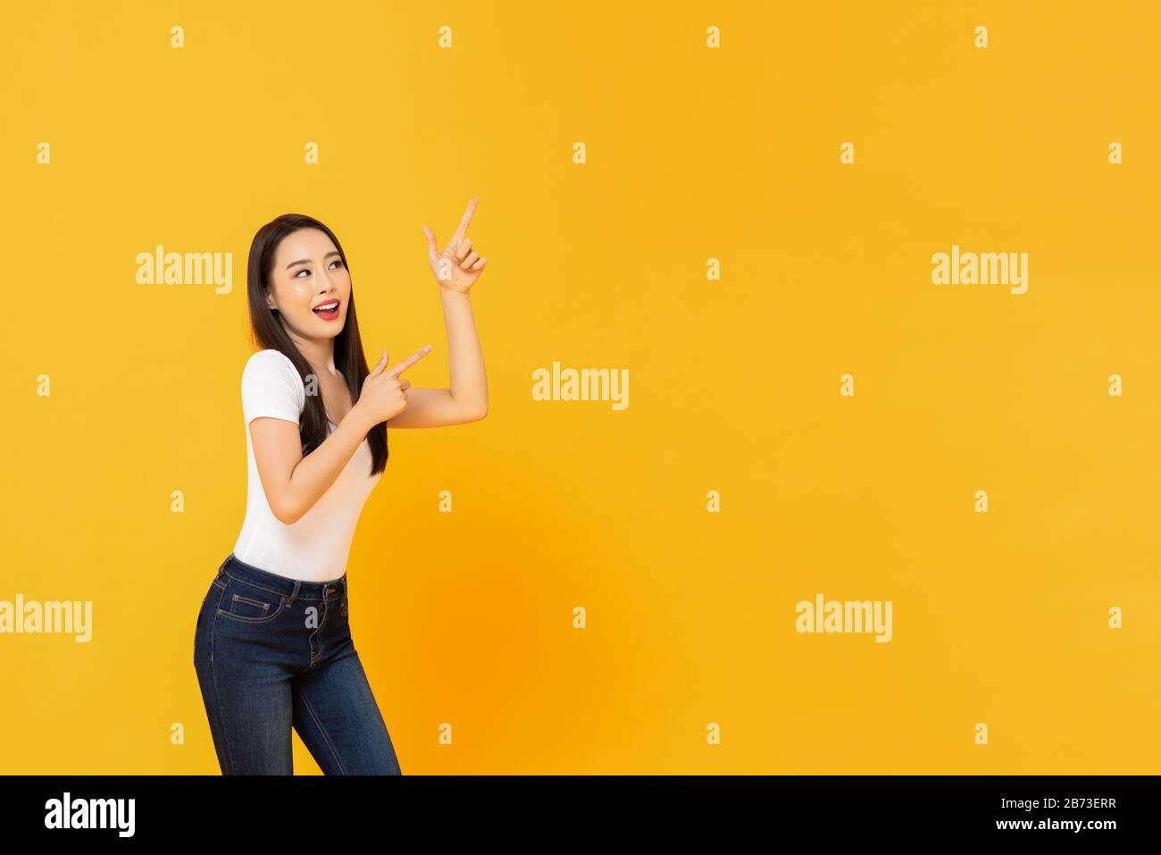 Portrait of smiling young beautiful Asian woman pointing up with her two fingers in blank space beside in yellow isolated studio background Stock Photo