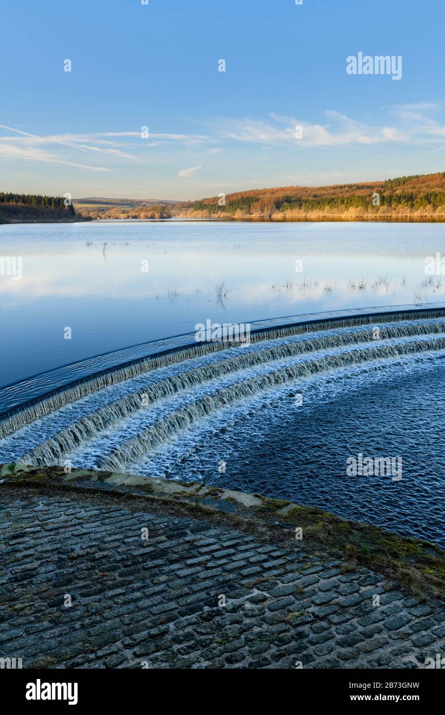 Water flowing over spillway from calm scenic tree-lined lake, under deep blue sky - Fewston Reservoir, Washburn Valley, North Yorkshire, England, UK. Stock Photo