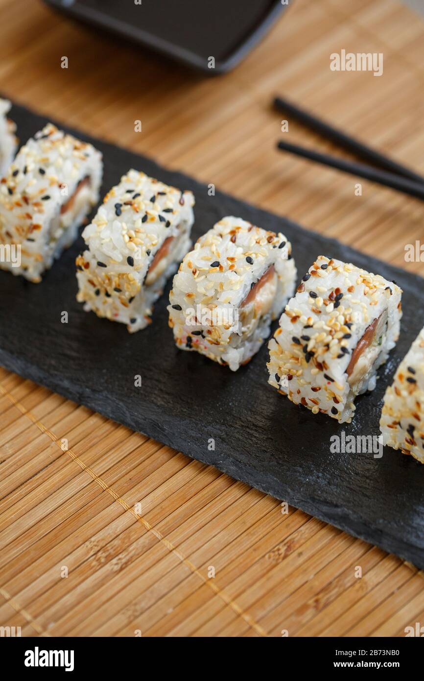 Uramaki served on a black slate with wooden black sticks and soy sauce. Japanese traditional food with bamboo service. Stock Photo