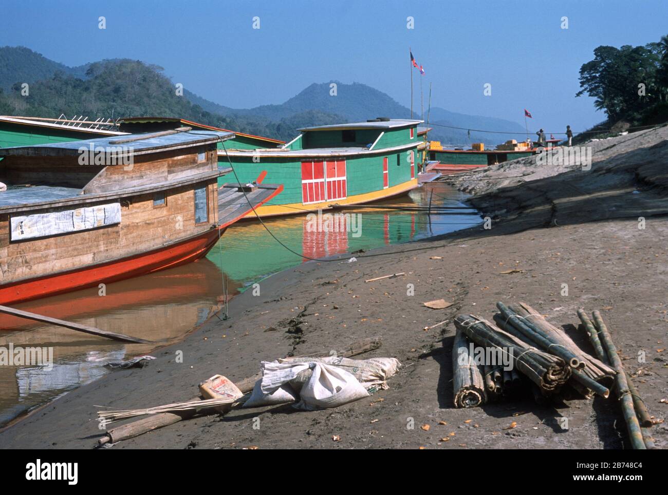 'Slow' boats tied up on the bank of the Mekong River in Laos. Lao flags fly from tall poles on each boat. Each boat has the traditional alter with offerings to the river on the foredeck. Cargo is waiting to be loaded on a jetty. Bright, red, green and yellow colours contrast with tree covered mountains and the blue sky behind. Stock Photo