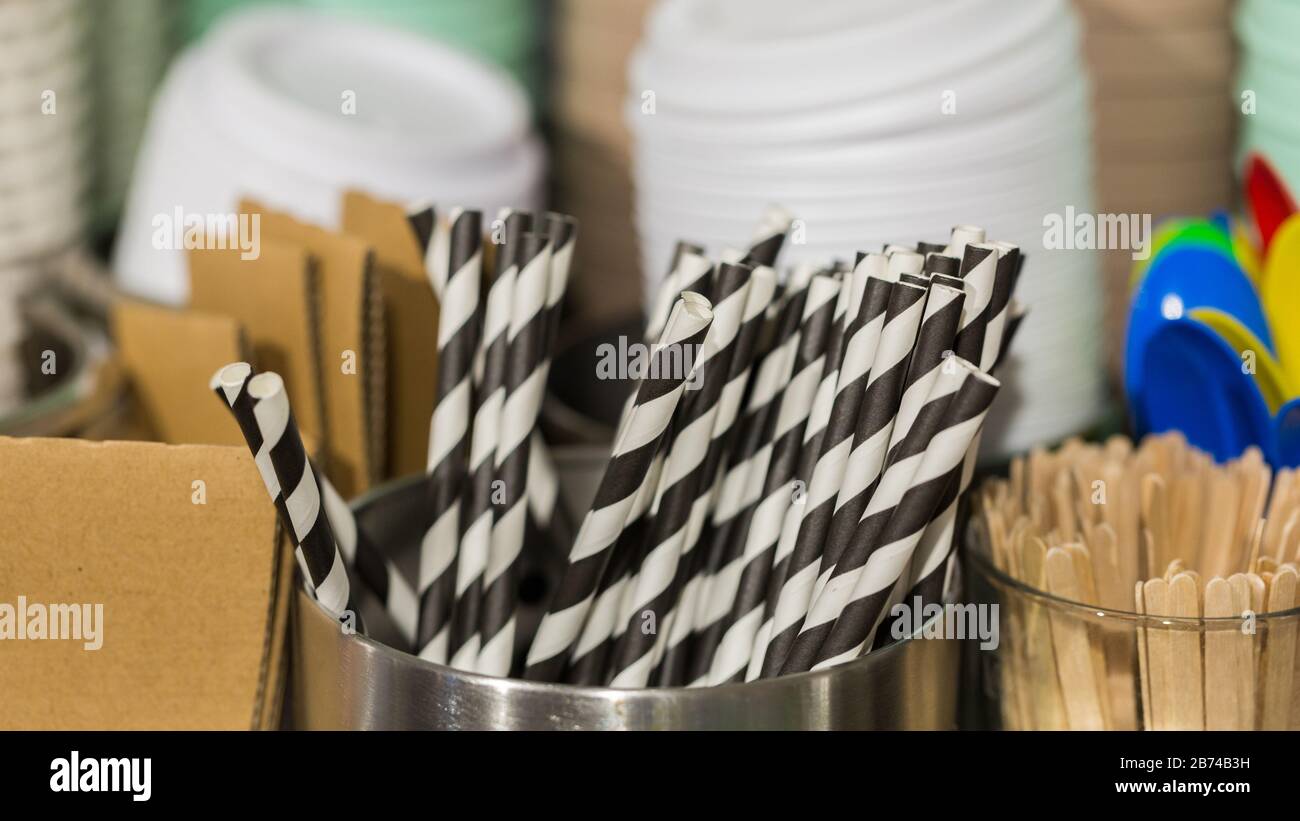Panorama with black & white striped drinking straws in a metallic bowl. On the right wooden sticks, in the back coffee cup covers. Coffee equipment. Stock Photo