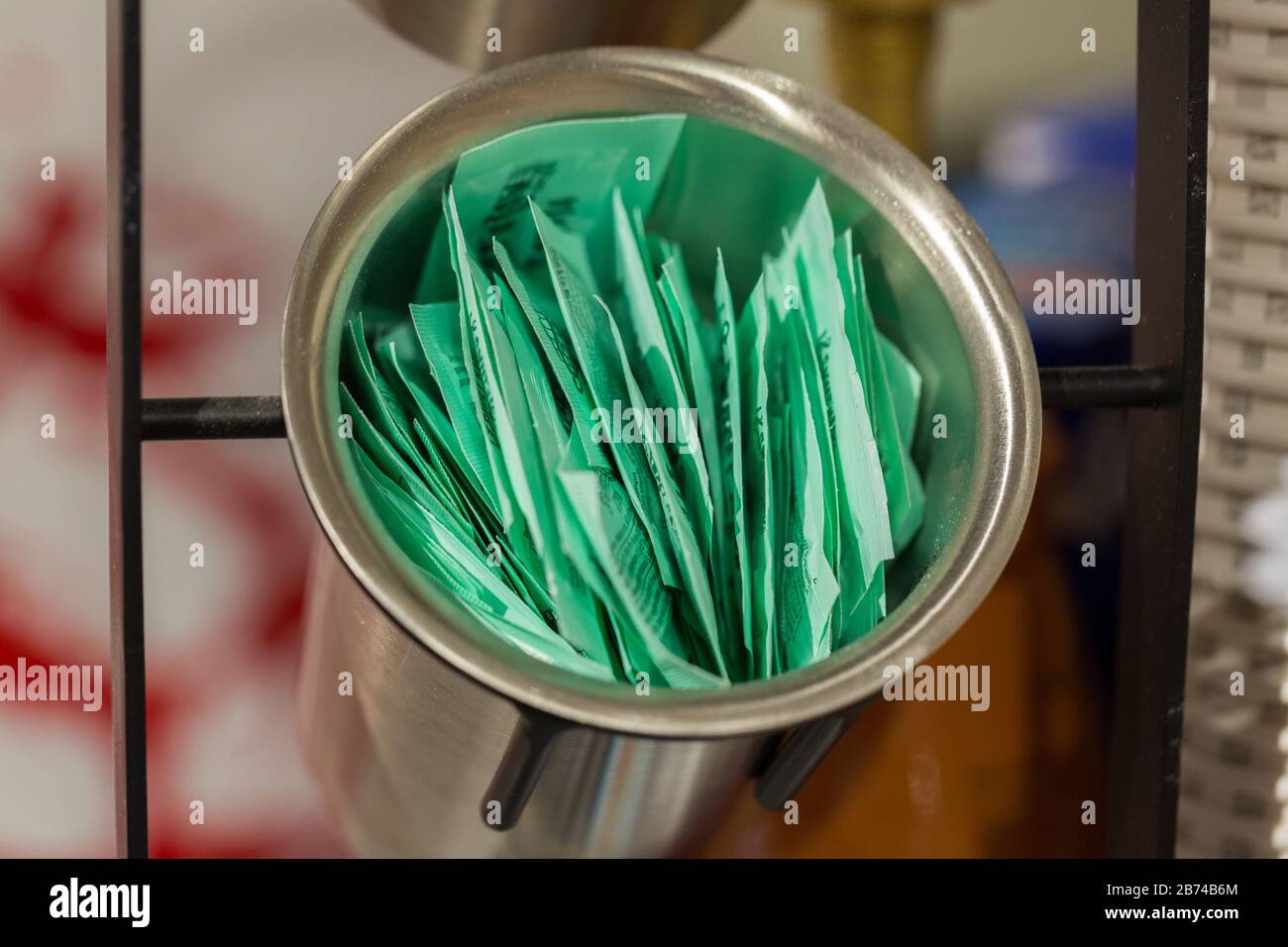 Sugar sachets in a metallic bowl. Many small, green paper bagsfilled with sugar. Neutral background, no logos. In a café. Stock Photo